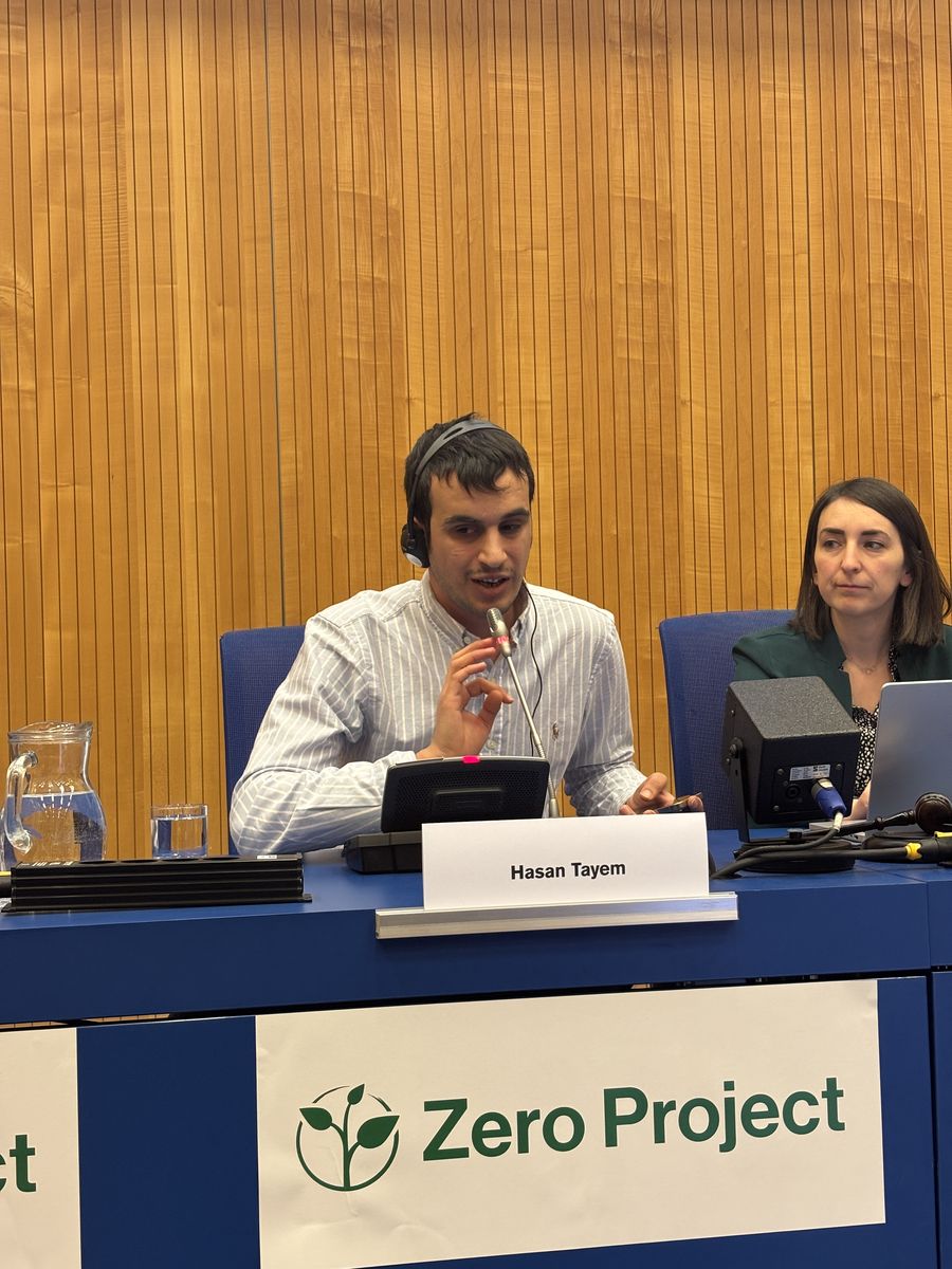 Hasan seated at a panel table with his nameplate at the Zero Project Conference at the United Nations Office in Vienna, with a fellow panellist beside him