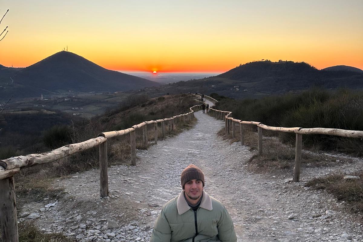 Hasan smiling at the camera while standing on a scenic mountain path at sunset, with hills and a glowing sky in the background