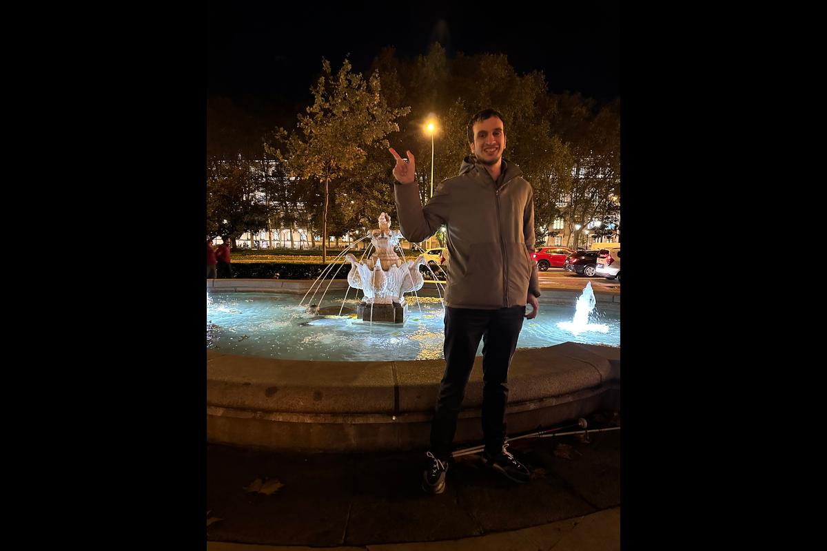 Hasan standing in front of a lit fountain in Madrid at night, smiling and making a peace sign with his right hand, with trees and city lights in the background