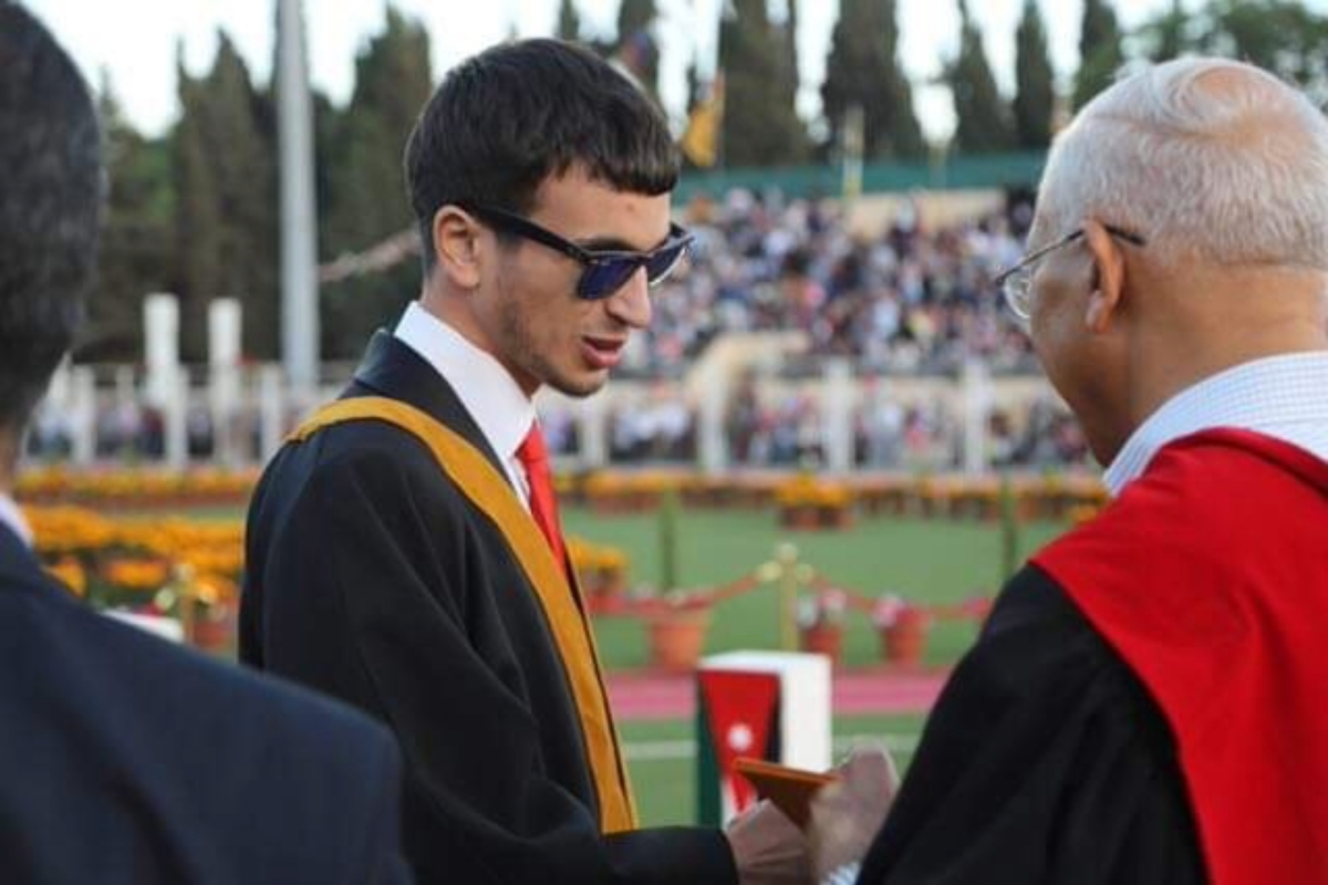 Hasan, wearing graduation robes and sunglasses, celebrates his bachelor's graduation outdoors by shaking hands with a distinguished guest